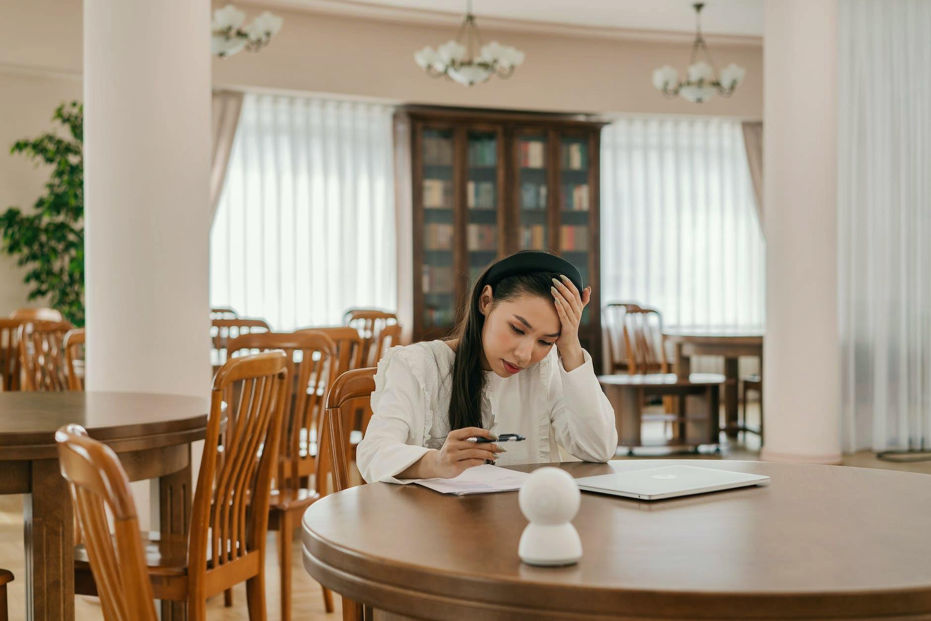 a woman in white long sleeves looking at the paper on the table with her hand on her head
